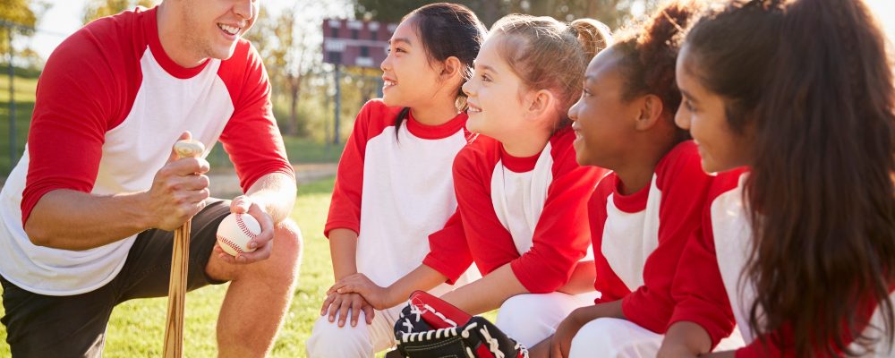 girl-baseball-team-kneeling-in-a-huddle-with-their-coach